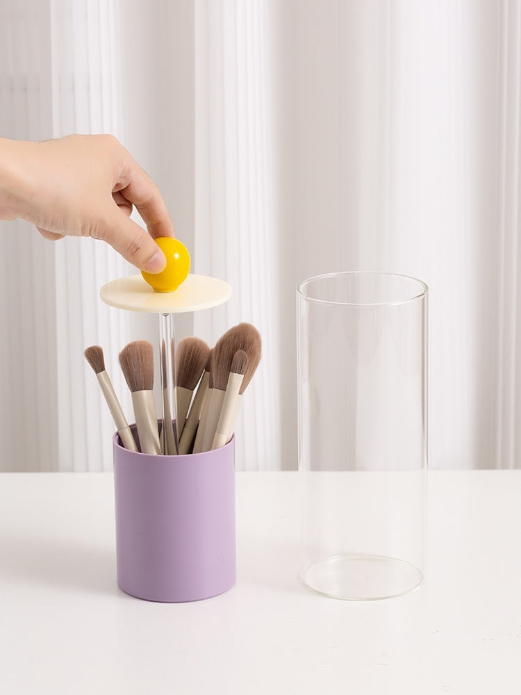 Hand placing a yellow ball into a small tower with makeup brushes inside, on a white surface.