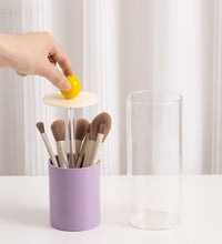 Hand placing a yellow ball into a small tower with makeup brushes inside, on a white surface.
