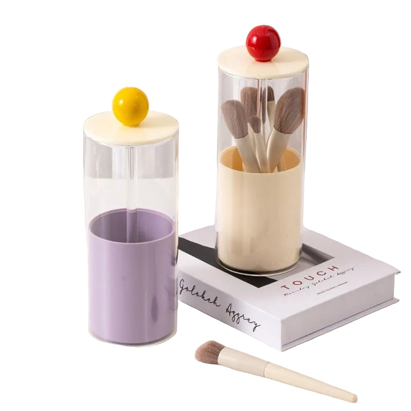 Makeup brush set in a cylindrical glass holder with a purple cup and a book on a white surface.