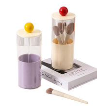 Makeup brush set in a cylindrical glass holder with a purple cup and a book on a white surface.