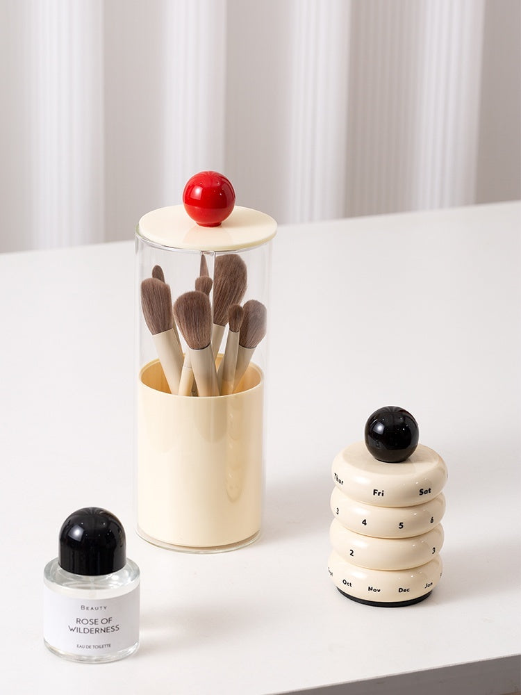 Set of makeup brushes in a cylindrical holder with a red ball on top, next to a small bottle and a stack of rings with numbers on a white surface.