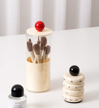 Set of makeup brushes in a cylindrical holder with a red ball on top, next to a small bottle and a stack of rings with numbers on a white surface.
